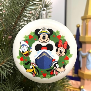 May include: A round white Christmas ornament featuring a green holly wreath graphic surrounding a blue cruise ship. The design includes cartoon character faces of Mickey Mouse, Minnie Mouse, Donald Duck, Pluto, and Goofy arranged around the ship.