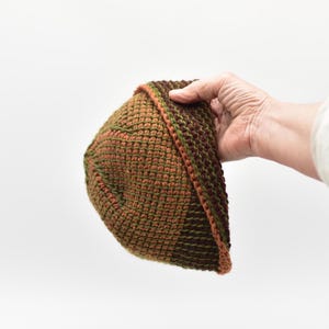 May include: A crochet beanie hat featuring a textured stitch pattern in alternating bands of dark brown, olive green, and terracotta orange yarn. The hat has a folded brim edge and a rounded crown, constructed with a dense, uniform yarn weave throughout.