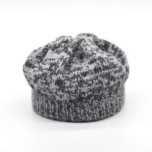 May include: A knitted beret featuring a variegated pattern of charcoal grey, light grey, and white yarn. The hat has a ribbed brim and a rounded, gathered crown, constructed with a textured knit stitch throughout the entire surface of the item.