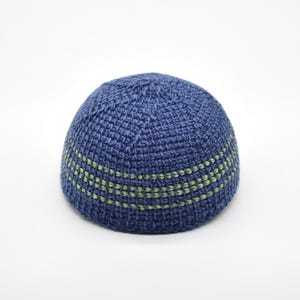 May include: A dome-shaped, hand-crocheted skullcap featuring a deep blue yarn base. The design includes two horizontal stripes of light green yarn woven into the texture, encircling the lower portion of the cap with a consistent pattern.