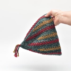May include: A triangular crochet hat featuring alternating stripes of dark green, mustard yellow, and deep red yarn. The hat has a textured knit pattern and includes a small decorative tassel made of matching yarn strands at the pointed tip.