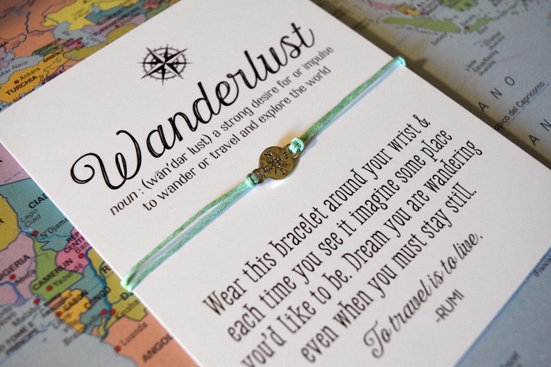 May include: A mint green bracelet with a compass charm, displayed on a card with the word "Wanderlust" and a definition. The card also features a quote about travel and exploration, set against a world map.