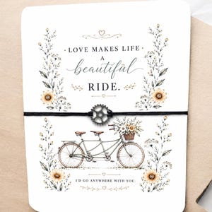 May include: A greeting card with a tandem bicycle illustration, a black string, and a metal gear charm. The card features the text "LOVE MAKES LIFE A beautiful RIDE. I'D GO ANYWHERE WITH YOU." Floral accents surround the bicycle.