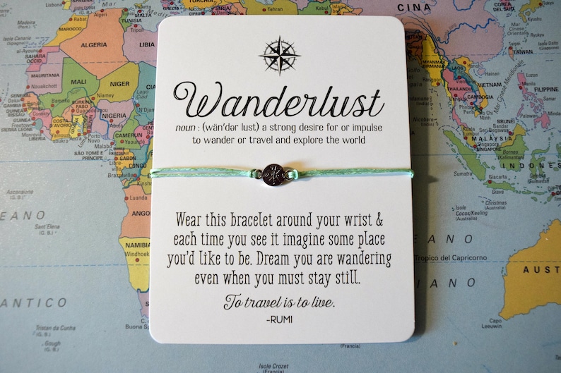 May include: A white card with the word "Wanderlust" and a definition, a compass graphic, and a bracelet. The bracelet is light green with a dark metal charm. The card is on a world map.