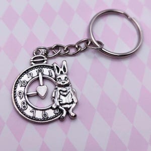 May include: Silver-toned keychain featuring a white rabbit with a pocket watch. The watch face has Roman numerals and a heart-shaped hand.