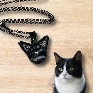 May include: A black wire-wrapped pendant necklace with a black and white stone and green accents. The pendant is shaped like a cat's head. A black and white cat is sitting on a wooden surface.