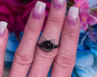 May include: A black wire-wrapped ring with a black gemstone in the shape of a cat's head. The ring is on a person's finger.