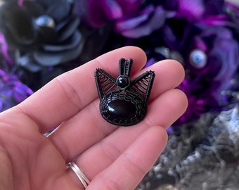 May include: A black wire-wrapped pendant with a black gemstone in the center. The pendant is in the shape of a cat's face.