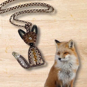 May include: A wire-wrapped fox pendant necklace with a brown chain. The pendant is made of copper wire and features a clear crystal in the center of the fox's body. The fox is sitting on a wooden surface.