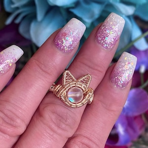 Könnte beinhalten: Ein goldfarbener, drahtgewickelter Ring mit einem schillernden weißen Edelstein. Der Ring wird an einer Hand mit rosafarbenem und weißem Glitzer-Nagellack getragen.