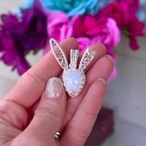 May include: A silver wire wrapped pendant featuring a pearlescent moonstone gemstone. The pendant is shaped like a rabbit with delicate wirework details.