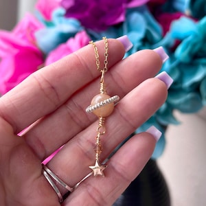 May include: A gold chain necklace with a peach-colored glass sphere pendant wrapped in gold wire. A gold star charm hangs from the sphere.