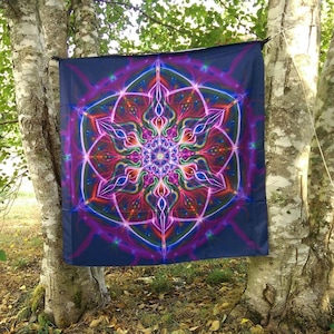 May include: A dark blue tapestry with a vibrant, symmetrical design in shades of purple, pink, green, and orange. The artwork features a central floral-like pattern with radiating lines and geometric shapes. The tapestry is hanging between two trees.