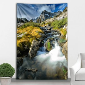 Puede incluir: Un tapiz con un paisaje de montaña con un arroyo que fluye. La imagen muestra una cordillera bajo un cielo azul con nubes. El arroyo fluye sobre rocas y está rodeado de vegetación verde y amarilla.