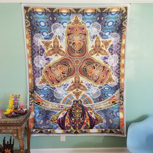 Peut inclure: Une grande tapisserie colorée avec un design symétrique complexe. L'œuvre présente une figure centrale avec un visage doré et des motifs complexes dans des tons de bleu, violet, orange et marron. La tapisserie est accrochée à un mur vert clair.