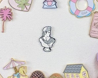 May include: A collection of enamel pins arranged on a white fabric stretched within a wooden embroidery hoop. The pins feature various designs, including a heart with a cannabis leaf, a bust, a house, and the word "DOLLHOUSE".