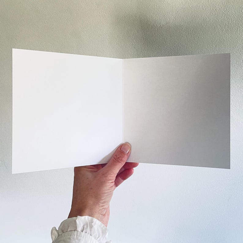 May include: A blank, open greeting card held by a person's hand. The card is white and has a smooth surface. The background is a light gray wall.
