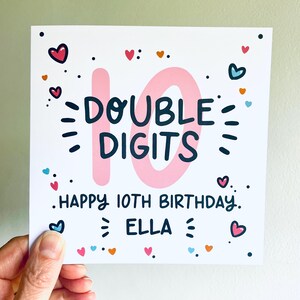 Op de afbeelding: Een roze en witte verjaardagskaart met het nummer 10 in roze en de tekst "DOUBLE DIGITS" in zwart. De kaart heeft ook de tekst "HAPPY 10TH BIRTHDAY" en "ELLA" in zwart. De kaart is versierd met kleurrijke harten.