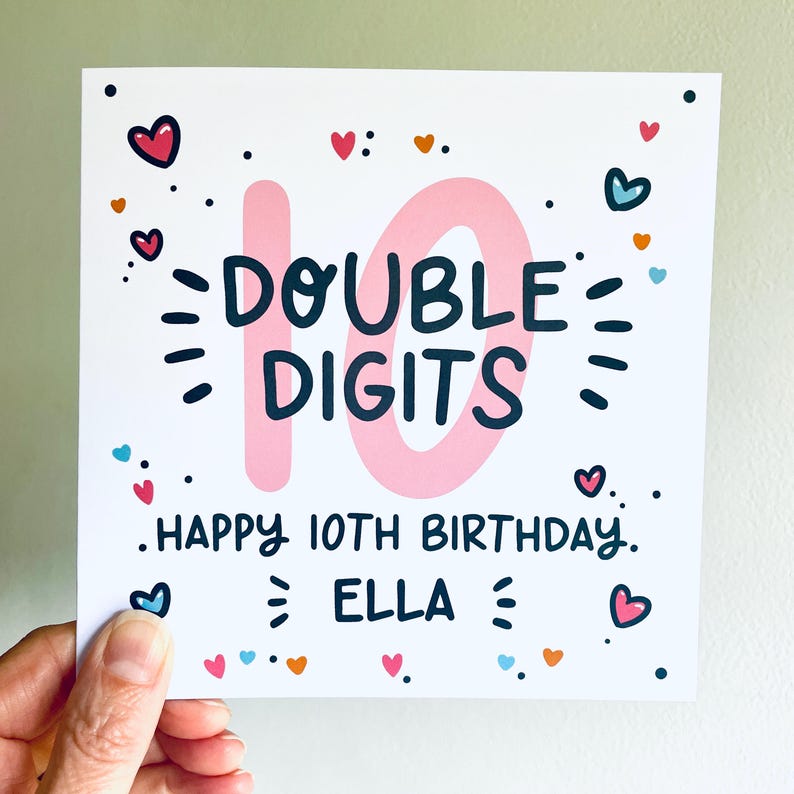 Op de afbeelding: Een roze en witte verjaardagskaart met het nummer 10 in roze en de tekst "DOUBLE DIGITS" in zwart. De kaart heeft ook de tekst "HAPPY 10TH BIRTHDAY" en "ELLA" in zwart. De kaart is versierd met kleurrijke harten.