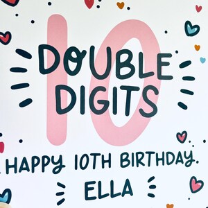 Op de afbeelding: Een roze en witte verjaardagskaart met de tekst "DOUBLE DIGITS" en "HAPPY 10TH BIRTHDAY. ELLA" in zwart. De kaart is versierd met hartjes en stippen.