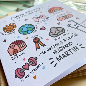 May include: A white greeting card featuring colorful hand-drawn illustrations and text. The card reads Happy Birthday to the one who is my best friend, is my world, is my rock, is my happy place, is my strength, and my gorgeous and lovely husband Martin.