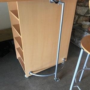 May include: A light wood desk with a metal bar attached to the underside. The bar is curved at the bottom and has a metal bracket on the side. The desk has shelves on the side.