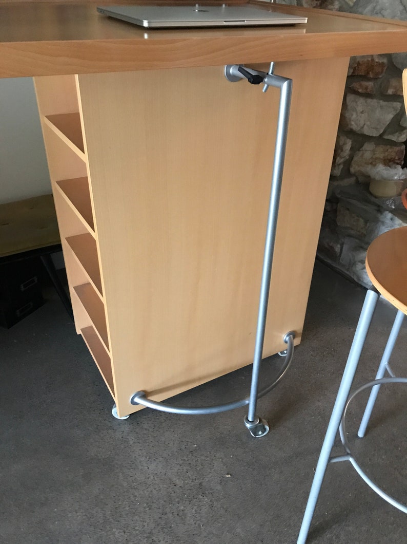 May include: A light wood desk with a metal bar attached to the underside. The bar is curved at the bottom and has a metal bracket on the side. The desk has shelves on the side.
