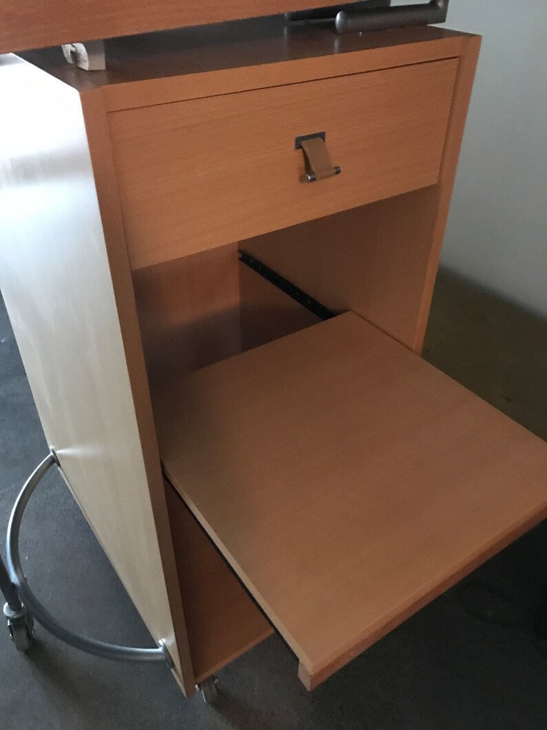 May include: A light wood mobile desk with a drawer and a pull-out shelf. The desk has a silver metal base with wheels.