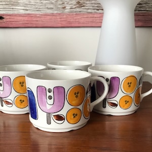 May include: Four white ceramic mugs with a colorful abstract design featuring purple, orange, blue, and brown accents. The mugs have a rounded shape and a handle on the right side.