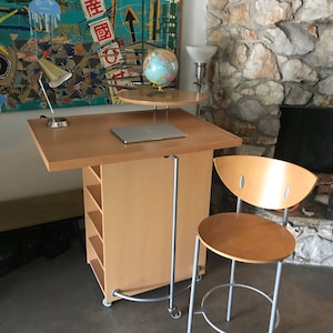 May include: A light wood rolling desk with a laptop, globe, and lamp on top. The desk has shelves and a metal bar for hanging items. A light wood chair with a metal frame is next to the desk.