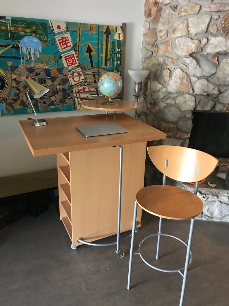 May include: A light wood rolling desk with a laptop, globe, and lamp on top. The desk has shelves and a metal bar for hanging items. A light wood chair with a metal frame is next to the desk.