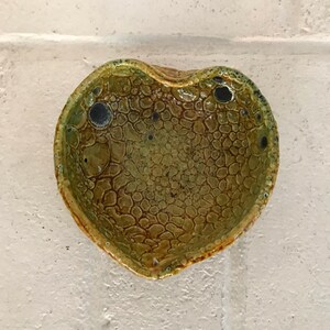 May include: A heart-shaped ceramic dish with a textured, green and brown glaze. The dish has a unique, organic shape and a speckled finish.