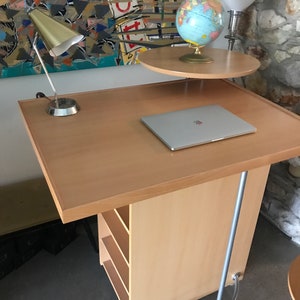 May include: A light wood rolling desk with a laptop, a lamp, and a globe on a smaller shelf. The desk has a silver metal base and four shelves.