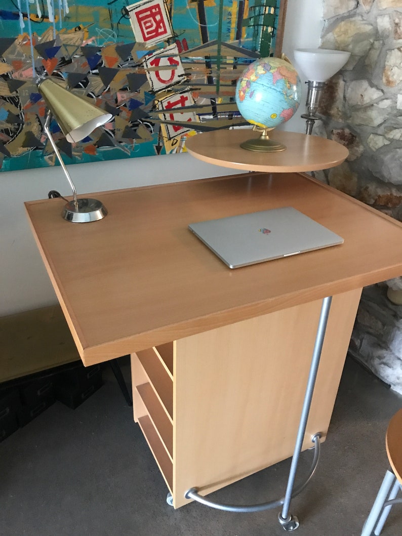 May include: A light wood rolling desk with a laptop, a lamp, and a globe on a smaller shelf. The desk has a silver metal base and four shelves.