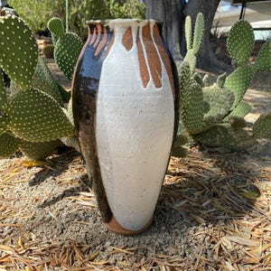 May include: A white ceramic vase with brown and black glaze. The vase has a rounded body and a narrow neck. It is sitting on a bed of brown mulch.