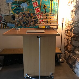 May include: A light wood mobile standing desk with a laptop on top. The desk has a silver metal frame and four wheels. The desk is in front of a colorful abstract painting with the text "畫圖" in red.