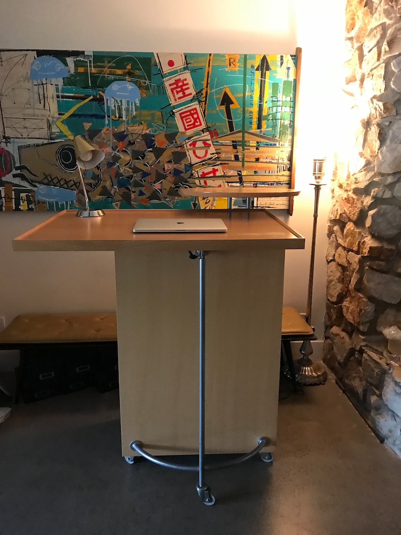 May include: A light wood mobile standing desk with a laptop on top. The desk has a silver metal frame and four wheels. The desk is in front of a colorful abstract painting with the text "畫圖" in red.