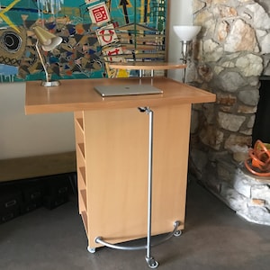 May include: A light wood standing desk with a laptop on top, a lamp on the left side, and a lamp on the right side. The desk has shelves and wheels. The desk is in front of a stone wall.