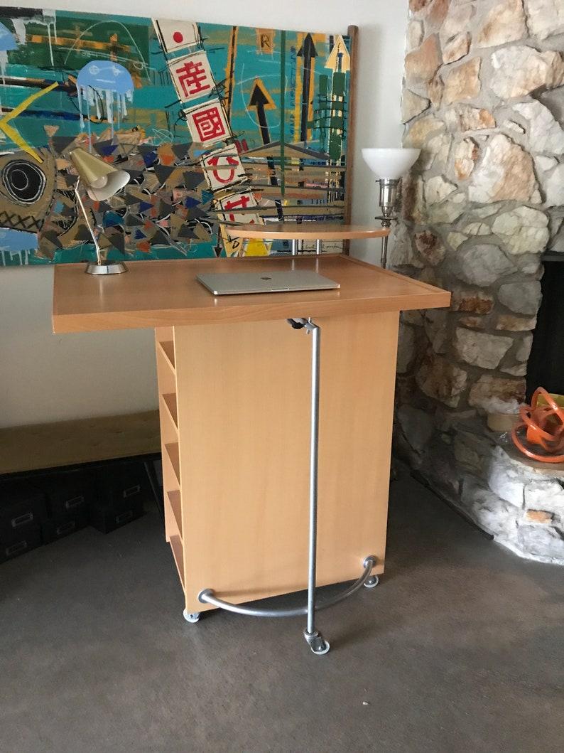 May include: A light wood standing desk with a laptop on top, a lamp on the left side, and a lamp on the right side. The desk has shelves and wheels. The desk is in front of a stone wall.