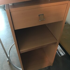 May include: A light wood mobile desk with a drawer and two shelves. The desk has wheels and a metal frame.