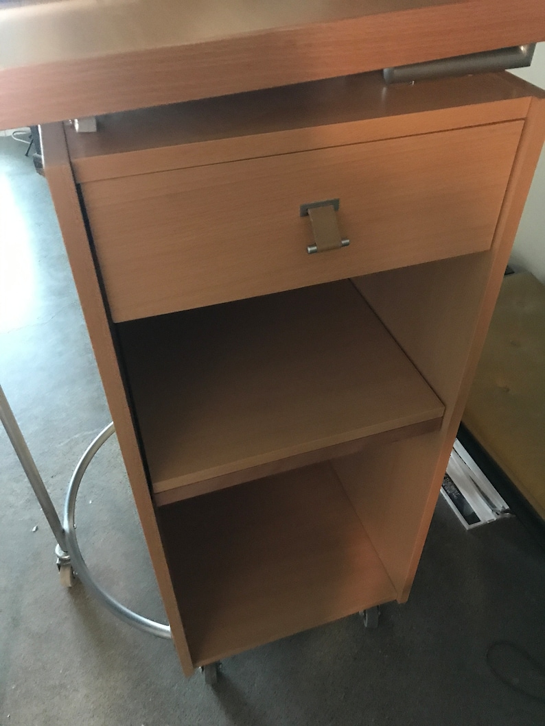 May include: A light wood mobile desk with a drawer and two shelves. The desk has wheels and a metal frame.