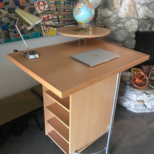 May include: A light wood rolling desk with a laptop, a lamp, and a globe on a smaller shelf. The desk has four shelves and is on wheels.