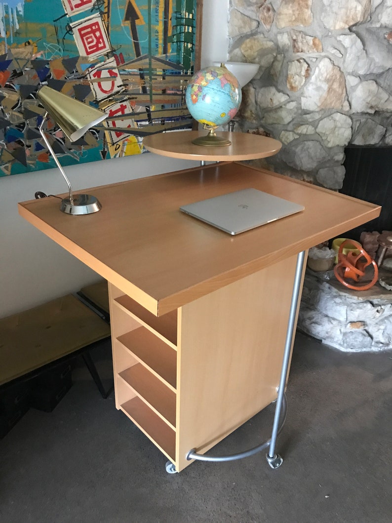May include: A light wood rolling desk with a laptop, a lamp, and a globe on a smaller shelf. The desk has four shelves and is on wheels.