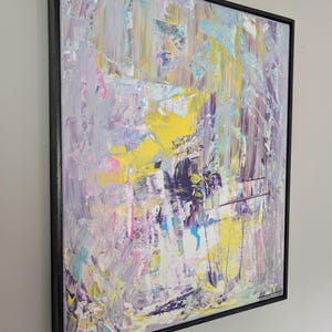 May include: Abstract art painting with a black frame. The artwork features a textured surface with a palette of colors including yellow, purple, blue, pink, and white. The brushstrokes create a dynamic and expressive composition.