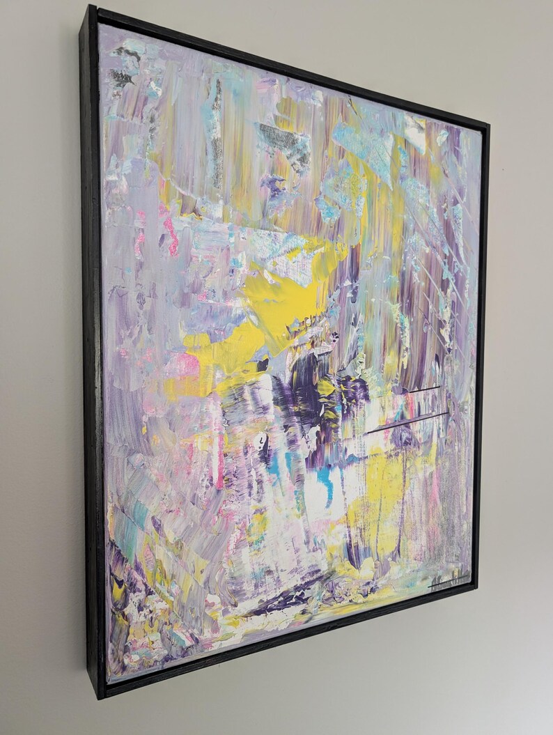 May include: Abstract art painting with a black frame. The artwork features a textured surface with a palette of colors including yellow, purple, blue, pink, and white. The brushstrokes create a dynamic and expressive composition.