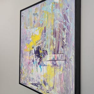 May include: Abstract painting with a black frame. The artwork features a textured surface with a blend of colors, including yellow, purple, blue, and pink. The brushstrokes create a dynamic and expressive composition.