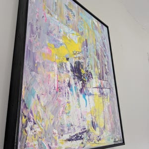 May include: Abstract art painting with a black frame. The artwork features a textured surface with a palette of colors including yellow, purple, pink, and blue. The brushstrokes are visible, creating a dynamic and expressive composition.