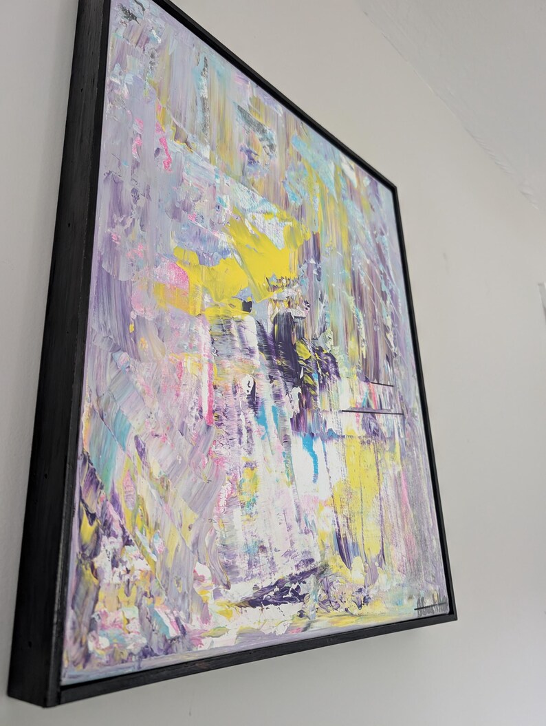 May include: Abstract art painting with a black frame. The artwork features a textured surface with a palette of colors including yellow, purple, pink, and blue. The brushstrokes are visible, creating a dynamic and expressive composition.