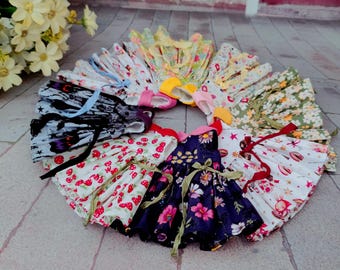 May include: A circular arrangement of colorful doll dresses. The dresses feature various floral and patterned designs, including strawberries, daisies, and stars. Each dress has a ribbon or bow, and the color palette includes red, blue, and yellow.
