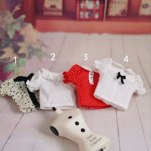 May include: Four doll-sized tops in different patterns and colors. The tops are white with black polka dots, white with a ruffled collar, red with white polka dots, and white with a black bow. The tops are arranged on a white surface with a doll-sized mannequin.